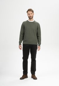 Groene trui van gebreid materiaal met geribde manchetten, gecombineerd met zwarte broek en bruine schoenen, staande in een minimalistische omgeving.