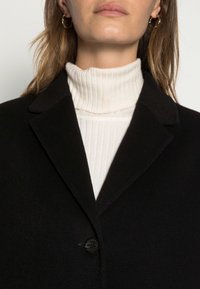 Cappotto di lana nero con collo strutturato, chiusura a un bottone, indossato sopra un dolcevita a coste color crema. Texture liscia, design minimalista.