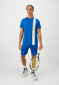 Modra atletska majica in kratke hlače z belim trakom, na katerem je napis "LACOSTE" v rumeni barvi. Drži tenis lopar in nosi bele superge.