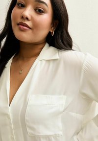 Vit, genomskinlig blus med krage, en bröstficka och slät textur. Modellen bär ett halsband med ett guldhjärta som hänger.