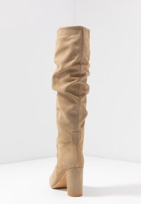 Bota beige por encima de la rodilla hecha de suave gamuza, con un diseño arrugado y un tacón grueso. Textura suave con punta redondeada.