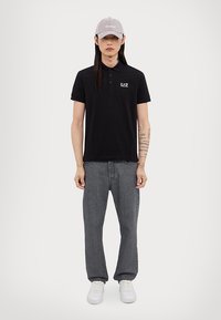 Čierna polokošeľa s krátkymi rukávmi a logom, spárovaná so sivými rovnými džínsami a bielymi teniskami. Sivá čiapka dopĺňa outfit.