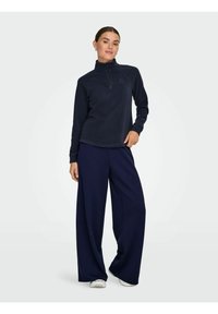 Tamno plavi polar pulover s visokim ovratnikom i pola patent zatvaračem, uparen s širokim tamnoplave hlače. Glatka tekstura s minimalnim brendingom.