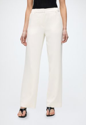 Kvinde iført højtaljede, vidde hvide bukser med sort ærmeløst top og sorte sandaler med åbne tæer mod en ensfarvet baggrund.