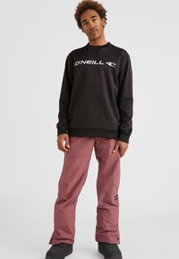 Zwarte sweater met wit logo, gecombineerd met roze skibroeken en zwarte laarzen. De sweater heeft een ronde halslijn en ribbelmanchetten.