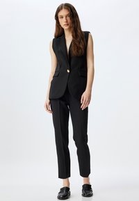 Blazer sans manches noir avec un col cranté et un bouton doré unique, associé à un pantalon noir ajusté et des mocassins noirs brillants.