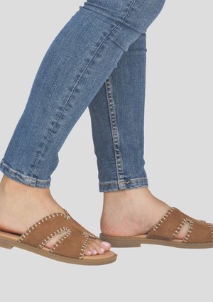 Pieds portant des sandales marron en daim à enfiler avec des coutures blanches et un jean en denim bleu sur un fond gris clair.