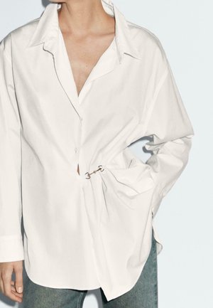 White oversized button-up shirt cinched at the waist with a metal clasp, worn with light blue jeans, model's hand in pocket, no visible face.