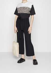 Mujer que lleva una camiseta de estampado de leopardo a bloques de color, pantalones negros de tiro alto con cinturón, zapatos de tacón negro y sostiene una bolsa de lona beige.