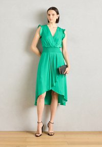Green, sleeveless dress with a wrap silhouette, smocked waist, and asymmetrical hem. Paired with a textured clutch and heeled sandals.