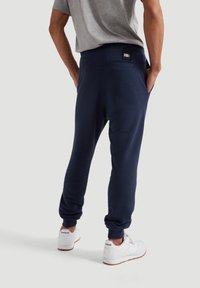 Navy blue sweatpants in soft fabric feature a relaxed fit, elastic waistband, and a back pocket detail. Paired with white sneakers.