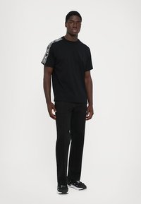 Μαύρη κοντομάνικη μπλούζα με λευκή ταινία λογότυπου στους ώμους, σε συνδυασμό με μαύρο tapered παντελόνι και μαύρα αθλητικά παπούτσια. Απλός, casual σχεδιασμός.