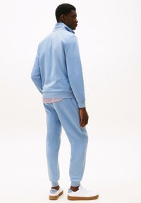 Ensemble de survêtement en polaire bleu clair avec un col montant, des poignets côtelés et des poches latérales, associé à des baskets blanches et une chemise rayée en dessous.