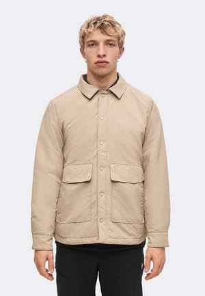 Jeune homme aux cheveux blonds courts et bouclés portant une veste beige boutonnée avec deux poches à rabat à l'avant et un pantalon noir sur un fond uni.