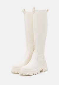 Bottes blanches montantes en matériau lisse avec une semelle épaisse, un bout rond et des tirants latéraux pour un enfilage facile.