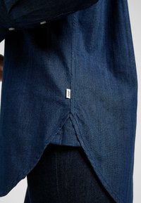 Jeansskjorta i mörkblått med en böjd fåll, med en liten märkesetikett. Tyget ser strukturerat och mjukt ut med framträdande sömdetaljer.