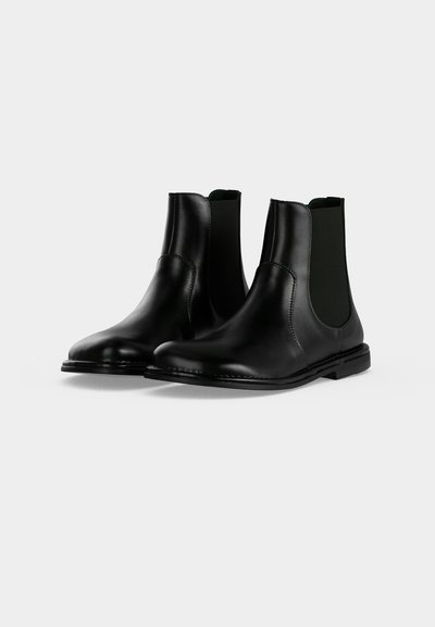 Paire de bottines en cuir noir avec panneaux latéraux élastiques et petits talons sur un fond clair uni.
