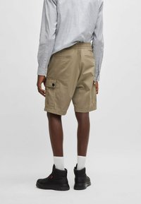 Pantaloni cargo beige con tasche laterali, regolatore di vita e tasche posteriori. Indossati con una camicia a righe blu e bianca e stivali neri.