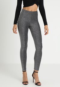 Hoge, glanzende zwarte leggings met een sprankelende textuur, een aansluitende pasvorm en een naadloos ontwerp, gecombineerd met zwarte, strappy hakken.