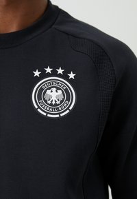 Crna sportska majica s logotipom Njemačkog nogometnog saveza koja sadrži orla i četiri zvjezdice na lijevom prsnom dijelu.