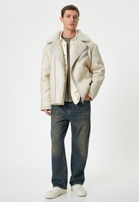Vit jacka med fårskinnsinsida, dragkedjefront, krämfärgad skjorta, grå t-shirt, mörkblå jeans och vita sneakers. Slät textur, avslappnad passform.