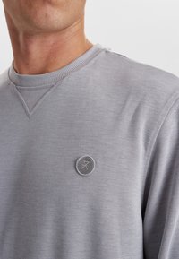Lys grå sweatshirt med ribbet rund hals, der har et lille cirkelformet logo på brystet. Blødt stof med en glat overflade.