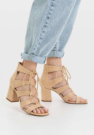 Pies calzados con sandalias de tacón bloque trenzadas de color beige claro con cordones, combinadas con vaqueros azul claro remangados sobre un fondo blanco.