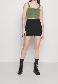 Crop top vert olive avec bordure à volants et fermeture à boutons, associé à un short taille haute noir et des bottines à talons noires.