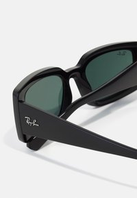Ochelari de soare dreptunghiulari negri cu lentile verzi. Au un cadru gros, design geometric și logo-ul argintiu "Ray-Ban" pe lateral.