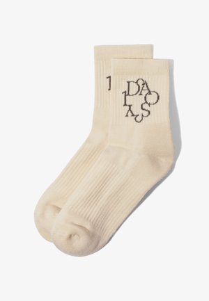 Chaussettes montantes côtelées couleur crème avec un motif de texte abstrait noir près du bord-côte, étalées à plat et superposées sur un fond blanc.