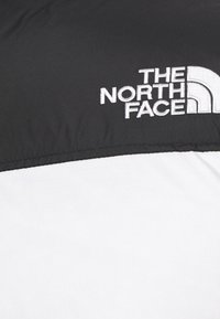 Veste matelassée avec la partie supérieure noire et la partie inférieure blanche, ornée du logo "The North Face" brodé en blanc sur la zone gauche de la poitrine.