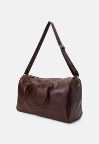 Καφέ δερμάτινη τσάντα ταξιδιού με στρογγυλό σχήμα, διπλές λαβές στην κορυφή και ρυθμιζόμενο λουρί ώμου. Διαθέτει ασημένια εξαρτήματα και κλείσιμο με φερμουάρ.