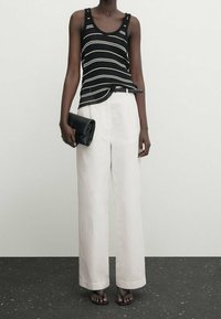 Top in maglia a righe bianche e nere con bottoni, abbinato a pantaloni bianchi larghi, tiene una pochette nera e indossa sandali piatti.
