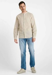 Lichtbeige overhemd met knopen van gestructureerde stof, met een opstaande kraag. Gecombineerd met lichtblauwe spijkerbroek en beige sneakers.