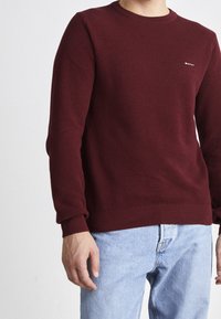 Bordeaux gebreide trui met ronde halslijn, ribgebreide manchetten en zoom. Voorzien van een gestructureerd patroon en een klein logo op de borst. Draagt bij lichte blauwe jeans.