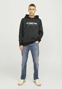 Zwarte hoodie met logo aan de voorkant, trekkoordkap en geribbelde manchetten, gecombineerd met blauwe jeans en crèmekleurige sneakers met groene en zwarte accenten.