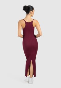 Vestido maxi de canalé burdeos con una abertura en el dobladillo y tirantes finos, confeccionado en tejido elástico. Llevado con zapatillas blancas sobre un fondo liso.