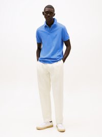 Kék pólóing pamutból, fehér nadrággal párosítva. A modell fehér sportcipőt visel bézs talppal, egy sima háttér előtt pózol.