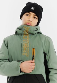 Jong persoon die de rits van een groen en zwart jack met een oranje "PROTEST" logo aanpast, terwijl hij een zwarte beanie draagt op een witte achtergrond.