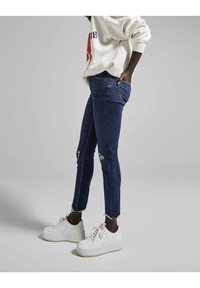 Jeans de mezclilla ajustados en azul oscuro con detalles desgastados y dobladillo sin terminar. Combinados con zapatillas deportivas de plataforma blancas. La modelo lleva una sudadera de color claro.
