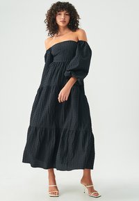 Svart, off-shoulder klänning med puffärm; texturerad tyg, lagerdesign och flytande silhuett. Bärs med vita sandaler med öppen tå.