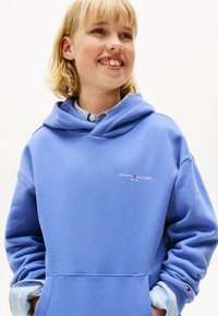 Glunderend kind in een blauwe Tommy Hilfiger hoodie over een gestreept shirt, met handen in de zakken, die lichtjes opzij kijkt tegen een witte achtergrond.