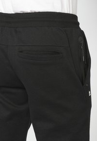  Sorte joggingbukser lavet af blødt materiale, med elastisk talje, sidelommer med lynlås og et lille logo på underbenet. Synlige stiklinger.