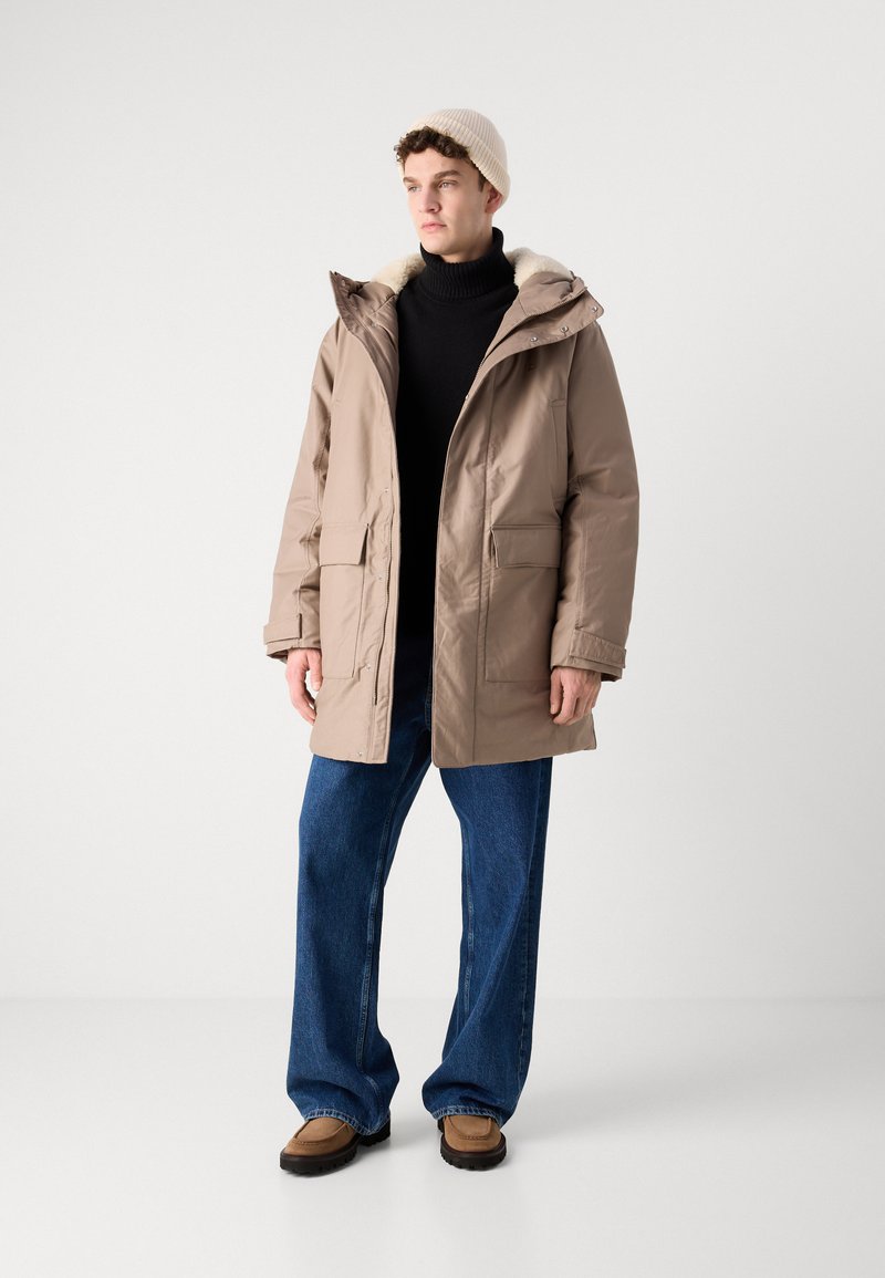 Beige gevoerde jas met capuchon, voorzien van een bontvoering, twee voorzakken, gecombineerd met een zwarte coltrui, blauwe wijde jeans en een paar bruine schoenen.