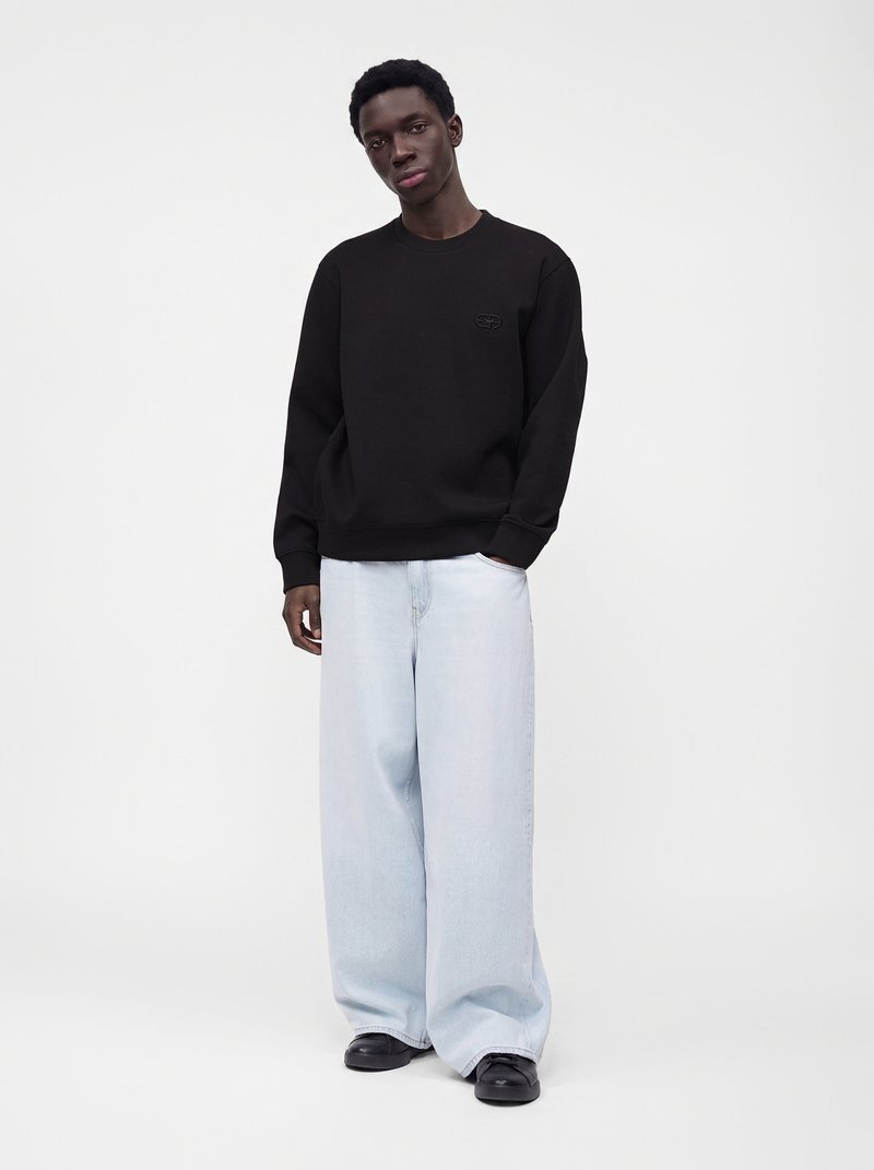 Man wearing black sweatshirt, light wide-leg jeans, and black shoes standing against plain white background.