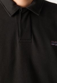 Černé polo tričko z texturované látky s límečkem a našitou logem v tmavě fialové barvě. Má knoflíkovou légu.