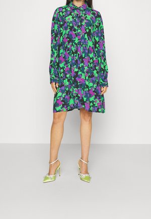 Woman wearing a loose, long-sleeve buttoned dress with green, purple, and black floral pattern, paired with lime green high heels with ankle straps.