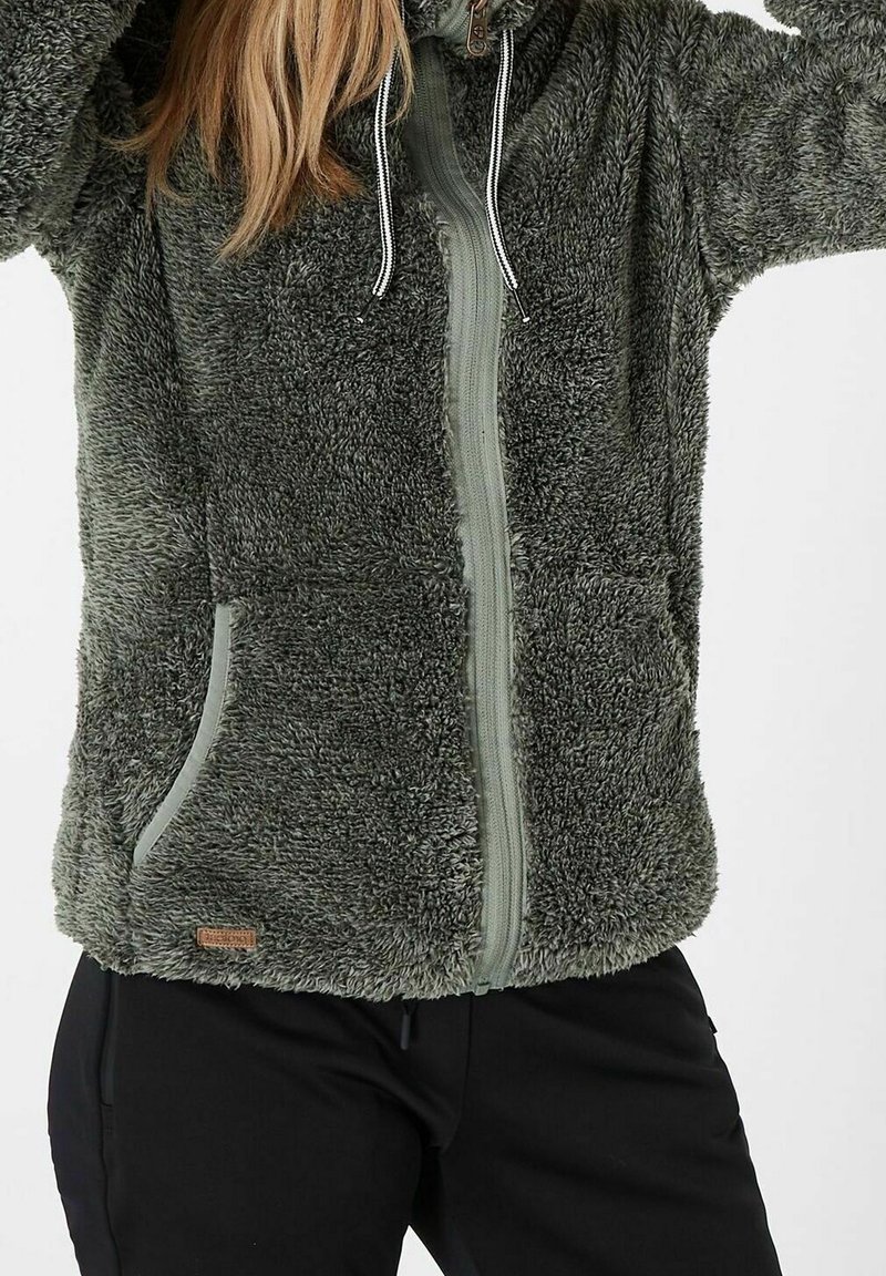 Femme portant une veste duveteuse gris foncé zippée avec poches et cordons, associée à un pantalon noir, sur un fond clair uni.