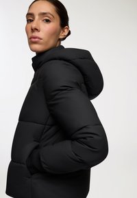 Czarna kurtka puchowa z kapturem, z pikowanym wzorem i bocznymi kieszeniami. Gładki, wyściełany materiał z prążkowanymi mankietami.