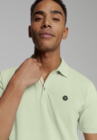 Lichtgroene polo met een kraagontwerp, korte mouwen en een hexagonaal zwart logo op de borst. Kenmerkt zich door een subtiele textuurstof.